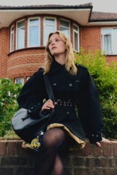 Model wearing a dark belted coat and holding a black shoulder bag while sitting on a brick wall in front of a brick house.