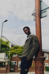 Man leaning against a brown street lamp pole wearing an olive green jacket and black trousers on a suburban street.