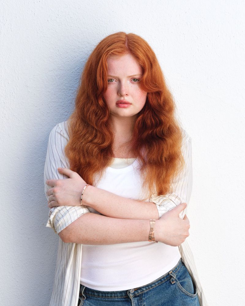 Young woman with long wavy red hair poses against a white wall in a casual white t-shirt, denim jeans, and cardigan.