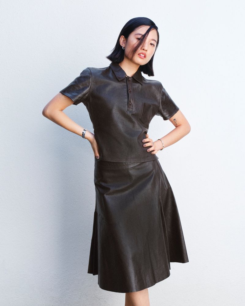 Woman in a dark leather skirt and matching polo shirt stands against a white wall in a studio, striking a fashionable pose.