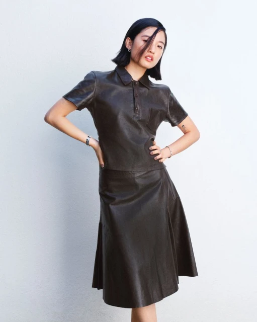 Woman in a dark leather skirt and matching polo shirt stands against a white wall in a studio, striking a fashionable pose.