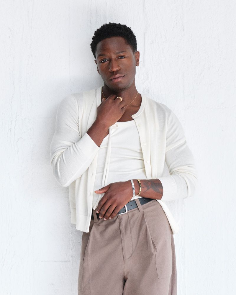 Adult male in a casual pose wearing a white cardigan, tank top, and brown slacks stands against a white wall in an indoor setting.