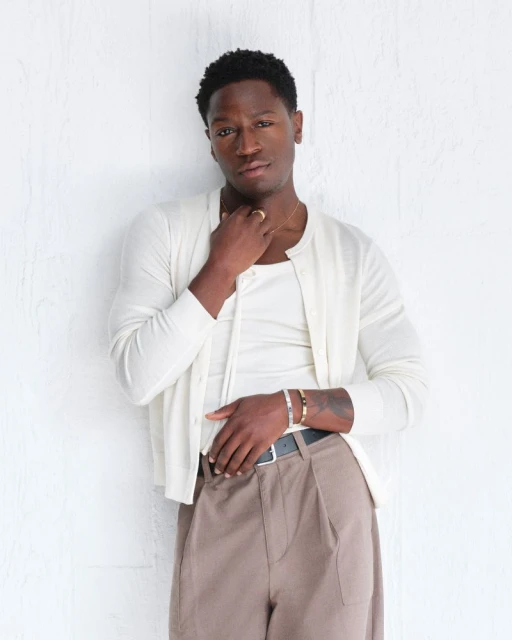 Adult male in a casual pose wearing a white cardigan, tank top, and brown slacks stands against a white wall in an indoor setting.