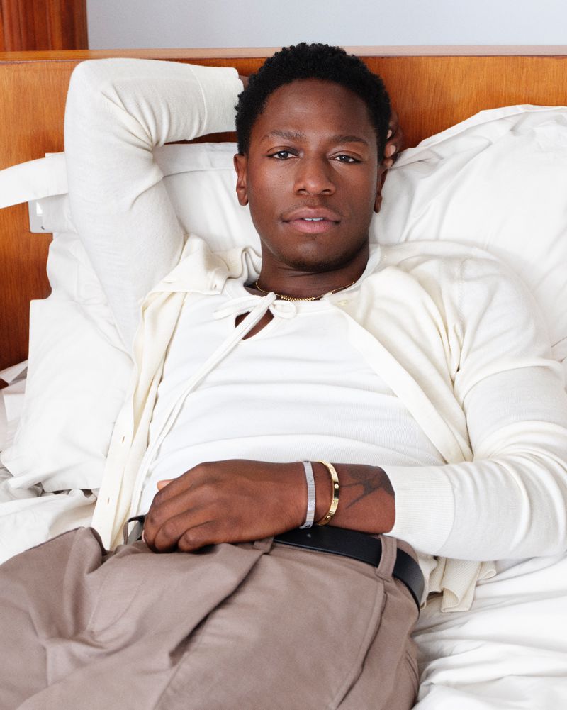 Portrait of an adult male relaxing in a bed with off-white linens, wearing a white shirt, cardigan, and tan pants, against a wooden headboard.
