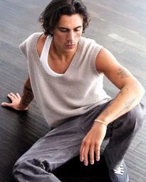 Man with arm tattoos wearing a beige knit sweater vest, white tank top, and grey trousers seated on a dark wood floor.