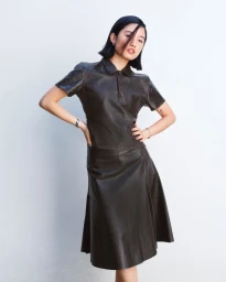 Woman with a dark bob haircut posing with hands on hips in a dark brown leather polo shirt and matching midi skirt against a white wall.