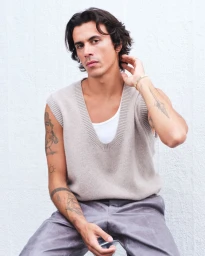 Man with wavy dark hair and arm tattoos wearing a beige knit sweater vest and gold bracelet against a white textured backdrop.
