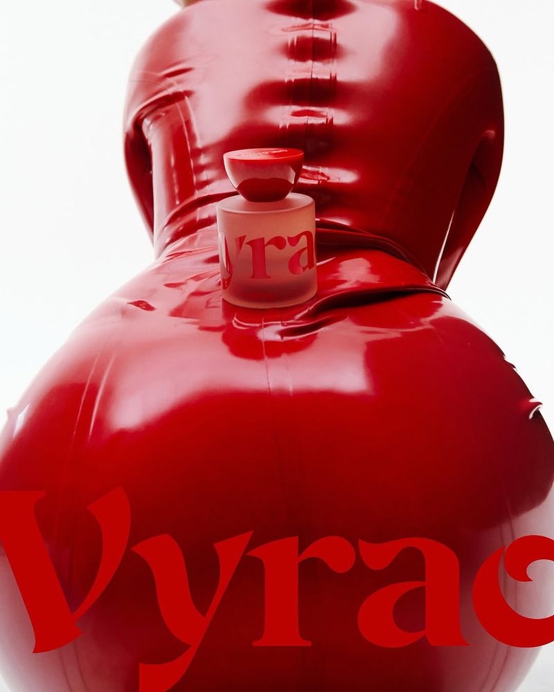 Red latex-clad figure presents a perfume bottle with 'Vyrao' text, set against a stark white background in a beauty and fashion context.