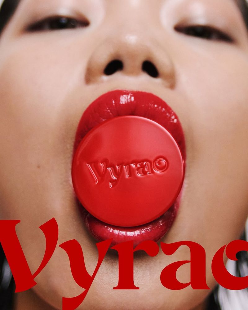 Close-up of a woman's face with red glossy lips holding a round red object with text in her mouth; beauty and cosmetics product shot.
