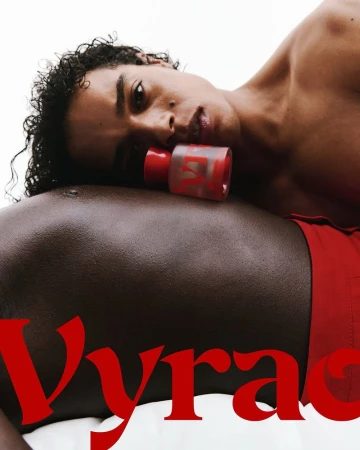 Two male models pose with a bottle of red-tinted perfume against a white backdrop in a close-up shot showcasing beauty and grooming.
