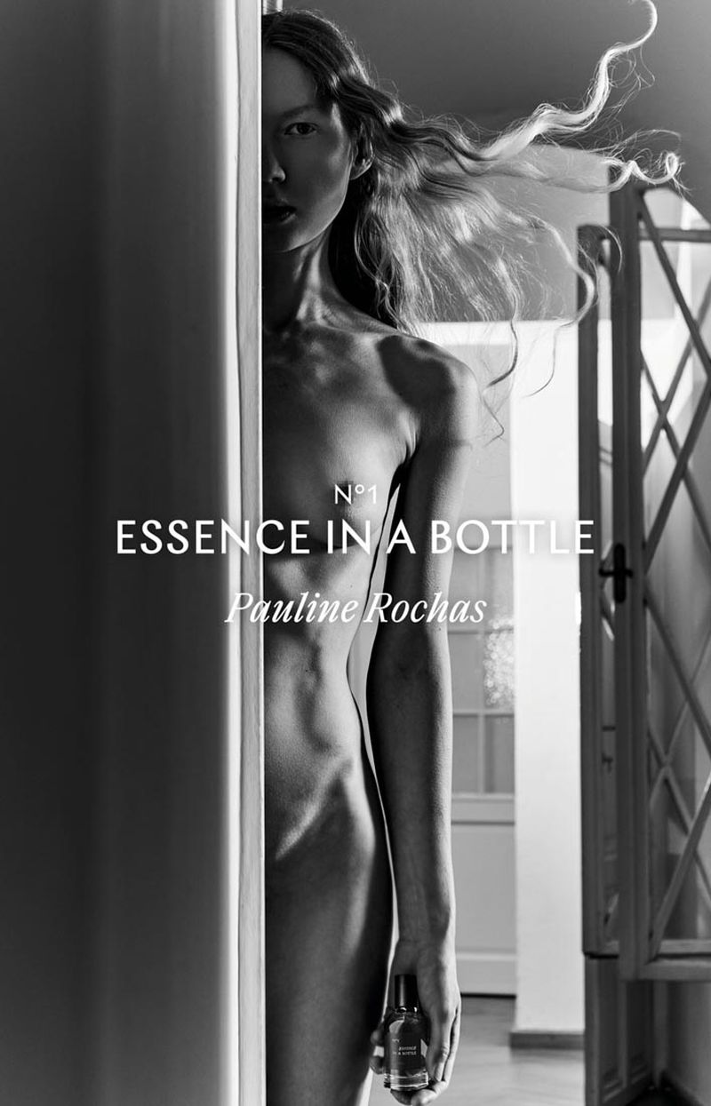 Black-and-white fashion portrait of a woman holding a perfume bottle, partially obscured by a doorway, against a bright, indoor background.