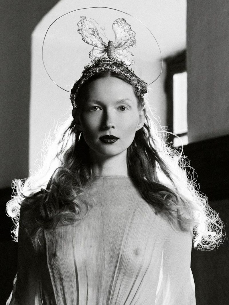 Monochrome portrait of a woman with a butterfly crown, long wavy hair, and dark lipstick, wearing a sheer gown in a studio setting.