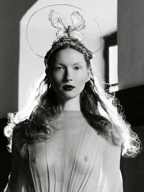 Monochrome portrait of a woman with a butterfly crown, long wavy hair, and dark lipstick, wearing a sheer gown in a studio setting.
