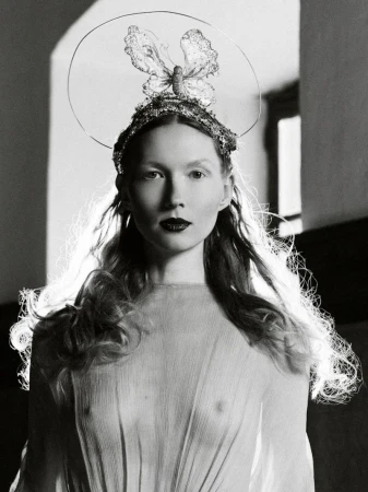 Monochrome portrait of a woman with a butterfly crown, long wavy hair, and dark lipstick, wearing a sheer gown in a studio setting.