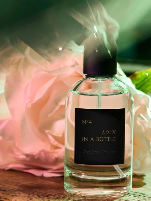 Close-up studio shot of a clear glass perfume bottle with black label on a wooden surface, with a large pink flower in the background.