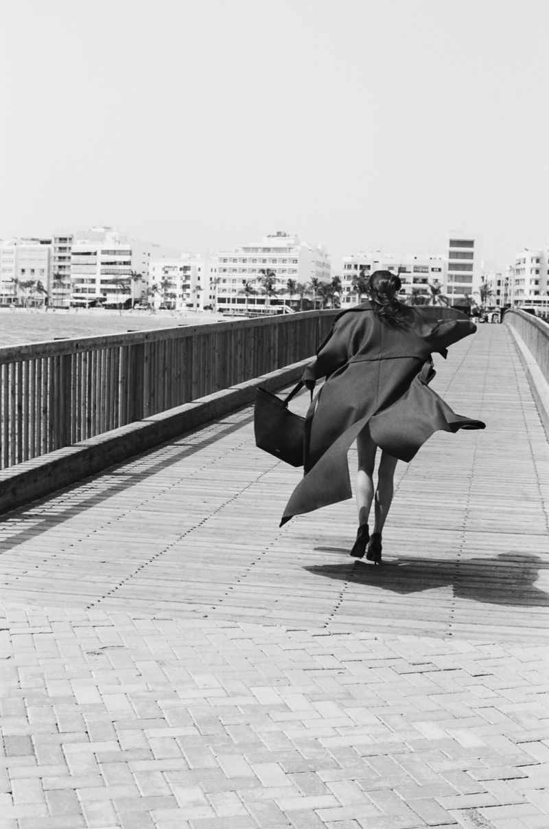 An adult female in a coat runs on a wooden bridge in an outdoor setting, with city buildings in the background, in monochrome.