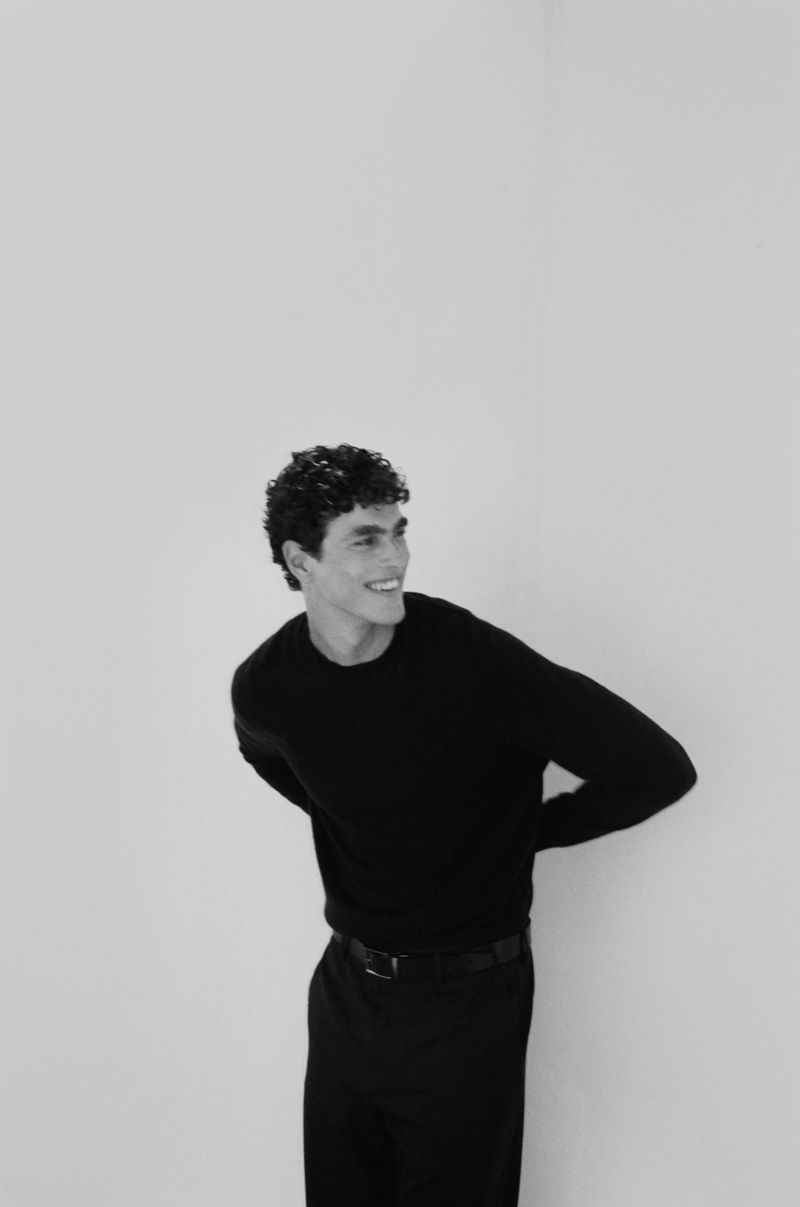Black and white portrait of a young adult male model with curly hair wearing a black sweater and black pants smiling in a photo studio.