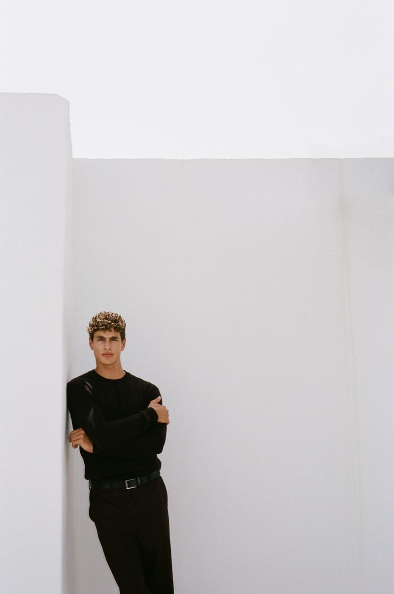Young adult male model with curly hair poses in black top and pants against white architectural wall in fashion-style studio shot.