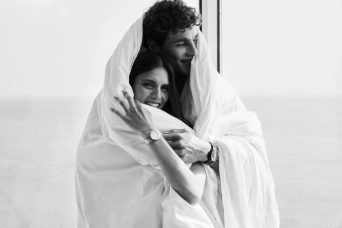 Black and white portrait of a smiling man and woman wrapped together in a large white blanket next to a window.