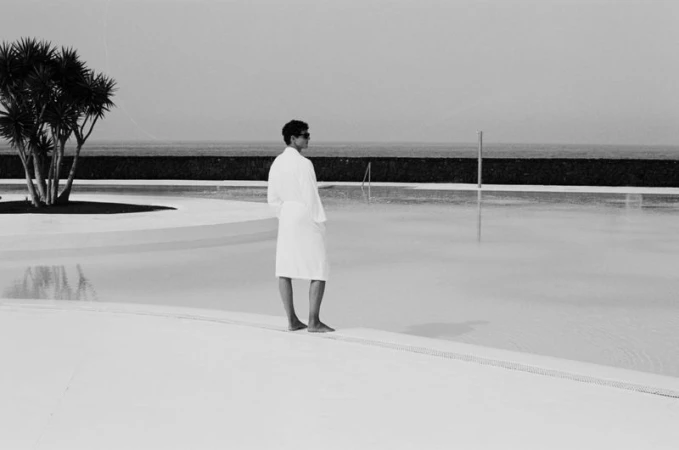 A man in a white bathrobe stands barefoot beside an outdoor pool overlooking the ocean horizon in black and white.
