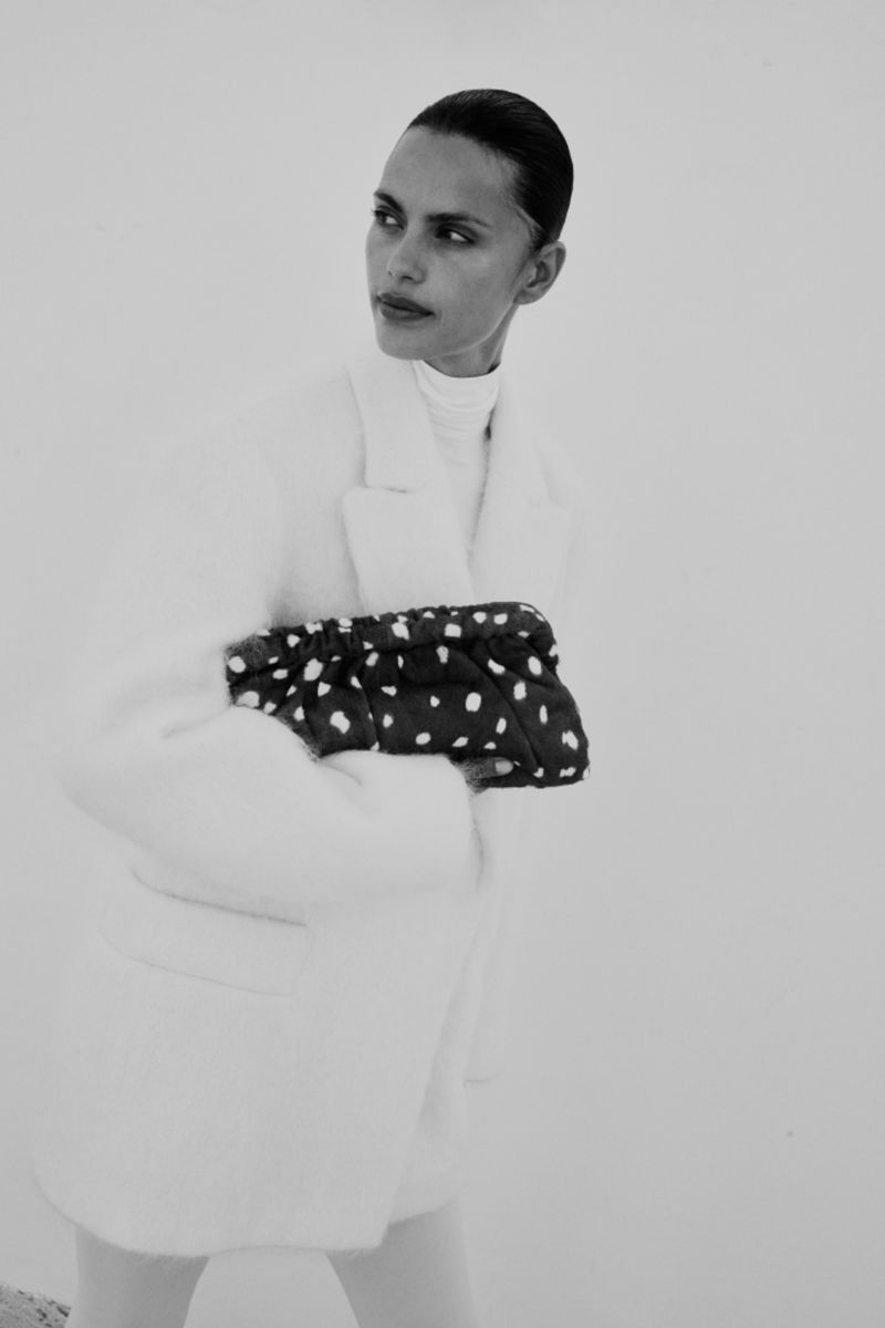 Monochrome fashion shot of a woman in a white coat holding a polka-dot clutch against a stark background, looking stylish.