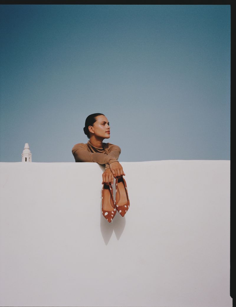 Woman in tan turtleneck poses with shoes against a white wall against a bright blue sky with a white building in the background.