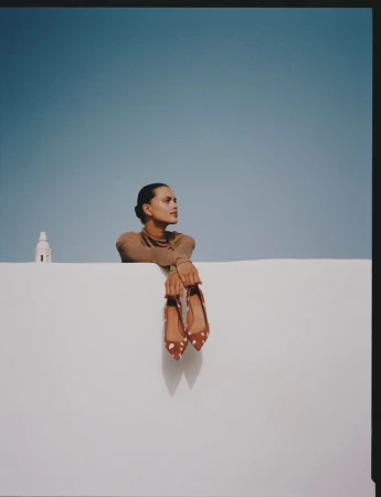 Woman in tan turtleneck poses with shoes against a white wall against a bright blue sky with a white building in the background.
