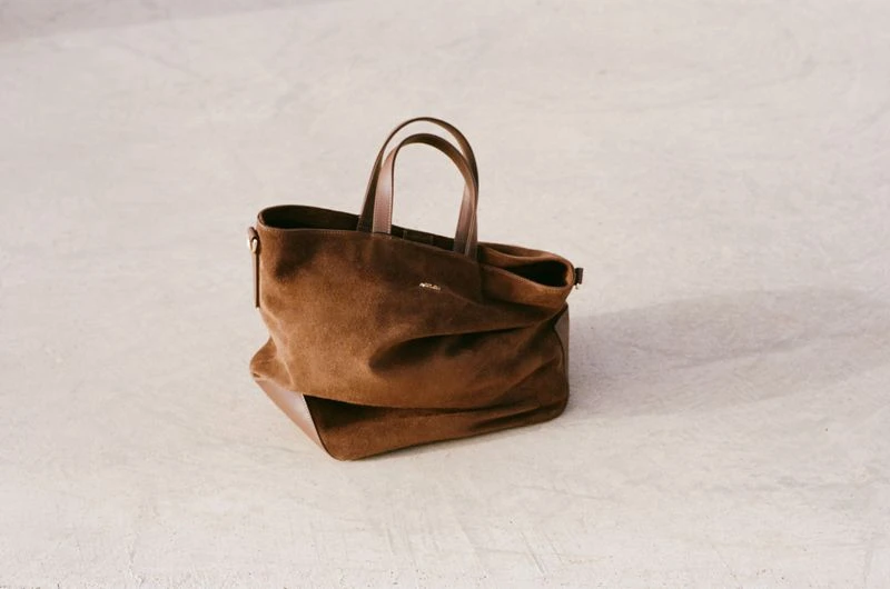 Brown suede tote bag with leather handles, isolated on a textured light-grey surface, showcasing fashion and design.