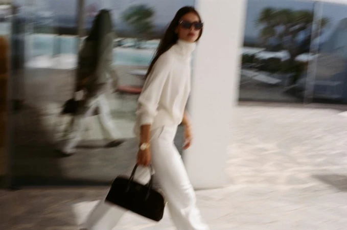 Model Myriam Tran in a white turtleneck sweater and white trousers carrying a black handbag outdoors in motion.