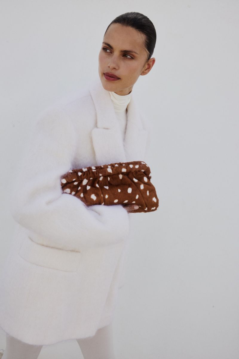 Fashionable woman in a white coat holds a brown clutch with white polka dots against a bright white background. Simple, stylish look.