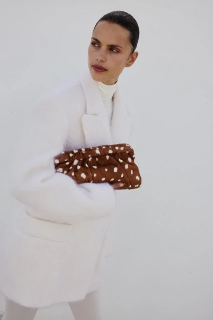 Fashionable woman in a white coat holds a brown clutch with white polka dots against a bright white background. Simple, stylish look.