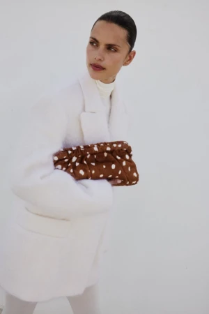Fashionable woman in a white coat holds a brown clutch with white polka dots against a bright white background. Simple, stylish look.