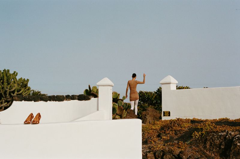 Adult female stands on a low rock wall in dress and tights near a white building with cacti, landscape view against pale sky.