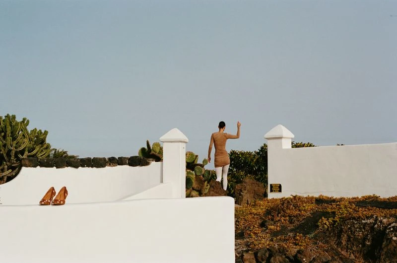 Adult female stands on a low rock wall in dress and tights near a white building with cacti, landscape view against pale sky.