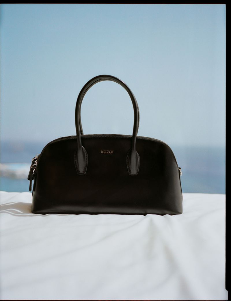 Elegant dark leather purse with dual top handles on a white textile against a blurred seascape and a clear blue sky background.