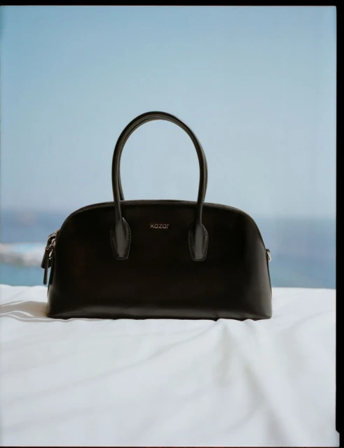 Elegant dark leather purse with dual top handles on a white textile against a blurred seascape and a clear blue sky background.