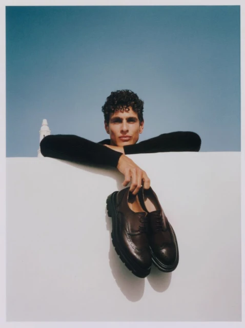 Male model leans on a white wall against a blue sky, holding a pair of brown leather dress shoes. Fashion editorial shot.
