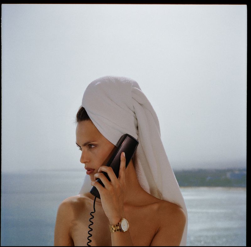 Woman with a white towel on her head talking on the phone in front of an ocean landscape on a sunny day, wearing gold watch.