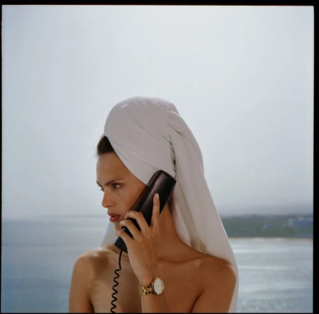 Woman with a white towel on her head talking on the phone in front of an ocean landscape on a sunny day, wearing gold watch.