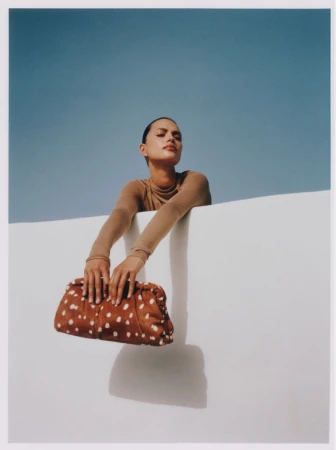 Adult woman with a tan turtleneck poses outdoors against a bright blue sky, holding a brown and white spotted purse against a white wall.
