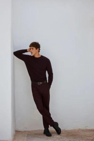 Full-length portrait of a young man in a dark long-sleeved shirt and trousers, leaning against a white corner in an indoor setting.