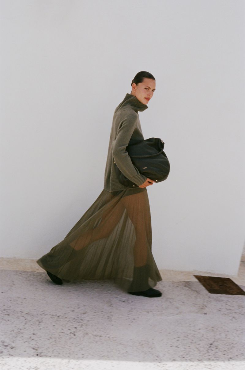 A woman wearing a green dress and carrying a black bag walks in an outdoor setting with a white wall as the backdrop.