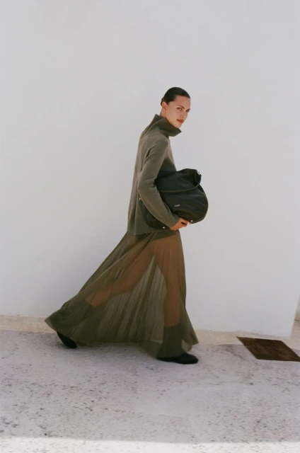 A woman wearing a green dress and carrying a black bag walks in an outdoor setting with a white wall as the backdrop.