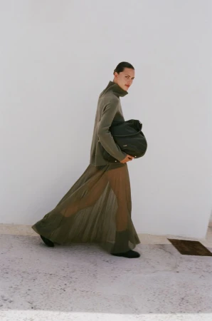 A woman wearing a green dress and carrying a black bag walks in an outdoor setting with a white wall as the backdrop.