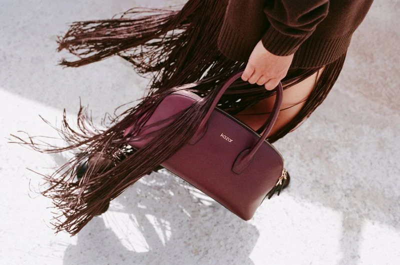 Burgundy leather handbag by Kazar is showcased with a brown sweater and fringed skirt on a light grey surface, fashion shot.