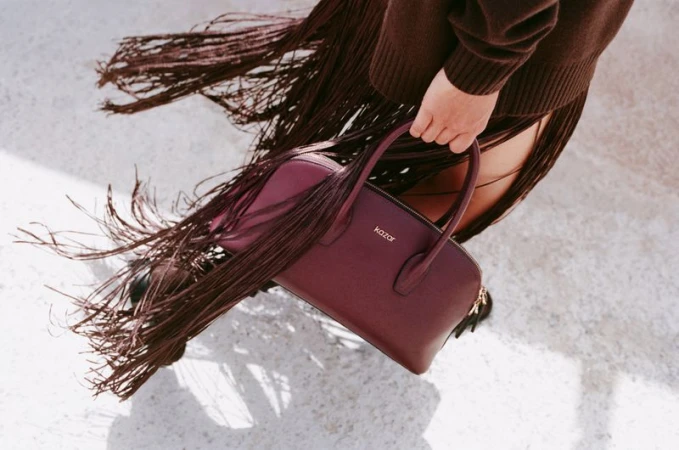 Burgundy leather handbag by Kazar is showcased with a brown sweater and fringed skirt on a light grey surface, fashion shot.