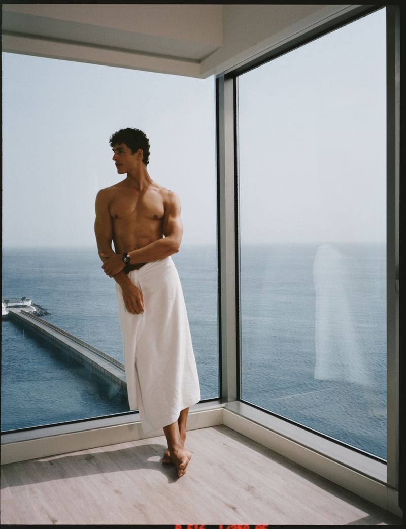 Athletic man wrapped in a towel stands in a corner with floor-to-ceiling windows overlooking the ocean on a bright, sunny day indoors.