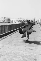 Model Myriam Tran seen from behind running along a wooden boardwalk in a long flowing coat and carrying a large tote bag.