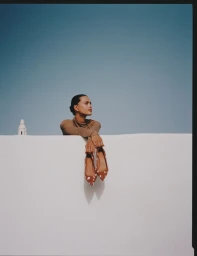 Model leaning on white wall holding patterned shoes against blue sky for Kazar A/W 25