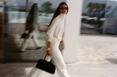 Model Myriam Tran in a white turtleneck sweater and white trousers carrying a black handbag outdoors in motion.